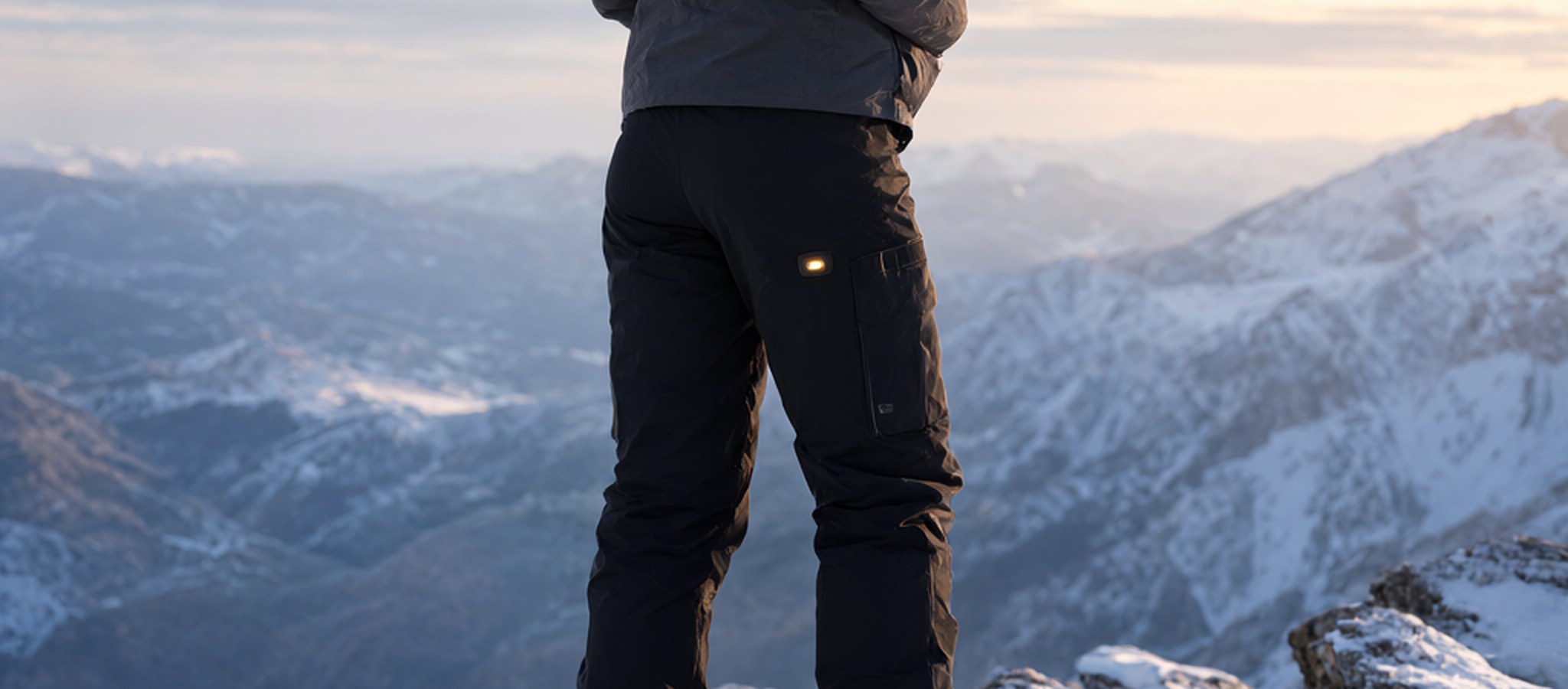 Woman in black heated softshell pants on a snowy mountain overlook at sunrise, face out of frame